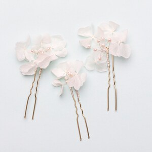 May include: Three gold hair pins with white hydrangea flower accents. The flowers are delicate and have a pearl-like finish.