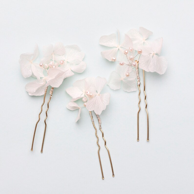 May include: Three gold hair pins with white hydrangea flower accents. The flowers are delicate and have a pearl-like finish.