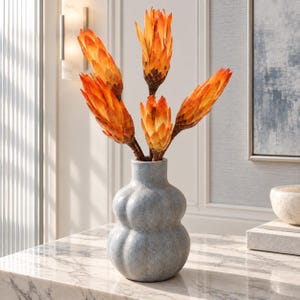 Puede incluir: Un jarrón de cerámica azul claro, texturizado y bulboso, que contiene cuatro flores de protea de color naranja vibrante. El jarrón tiene un cuello estrecho y un cuerpo redondeado con varios lóbulos. Las flores tienen pétalos puntiagudos.