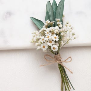 Könnte beinhalten: Ein kleiner Strauß weißer Blüten mit gelben Zentren und grünen Blättern, gebunden mit Bindfaden. Das Arrangement befindet sich vor einem weißen Hintergrund und erzeugt eine saubere und natürliche Ästhetik. Ideal für Hochzeiten oder besondere Anlässe.
