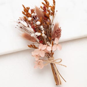 Könnte beinhalten: Ein kleiner Strauß aus getrockneten Blumen, mit Bindfaden gebunden. Die Anordnung weist eine Vielzahl von Texturen und Farben auf, darunter Orange, Weiß und Rosa. Der Strauß ist im natürlichen, rustikalen Stil gehalten, ideal für die Inneneinrichtung.
