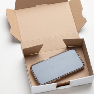 May include: A smartphone with a gold-colored case rests inside a white and brown cardboard box. The box is open, revealing the phone's screen. The box is on a white surface.