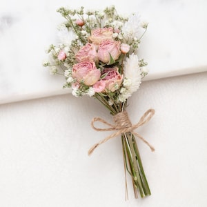 Könnte beinhalten: Ein Ansteckblümchen mit getrockneten rosa Rosen, weißem Schleierkraut und weißem Hasenschwanzgras. Die Stiele sind gebündelt und mit einer naturfarbenen braunen Schnur befestigt, die zu einer kleinen Schleife an der Basis gebunden ist.