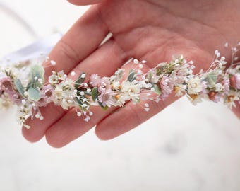 May include: A delicate floral crown featuring a variety of small, dried flowers in shades of white, pink, and green. The crown is held in a person's hand, showcasing its intricate design and natural aesthetic. The crown is perfect for weddings or special occasions.
