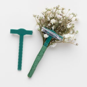 Könnte beinhalten: Eine grüne Blumennadel mit silberner Nadelrückseite. Die Nadel ist an einem Strauß weißer Baby's Breath Blumen befestigt.