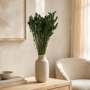 May include: A tall, cylindrical, matte beige ceramic vase holding a dense arrangement of slender, dark green dried grass stalks with small, textured seed heads. The vase has a smooth, uniform surface and a narrow neck, standing upright.