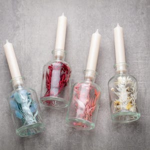 May include: Four clear glass bottles with white taper candles and decorative fillings. The fillings include blue, red, and white dried flowers. The bottles are arranged on a gray surface.
