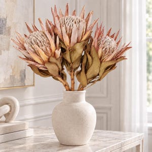 Könnte beinhalten: Ein Strauß aus drei großen, getrockneten Protea-Blüten mit staubig rosa, spitzen Blütenblättern und strukturierten, hellen Mitten. Die Blumen stehen in einer runden, cremeweißen Keramikvase mit einer matten, leicht rauen und gesprenkelten Oberfläche.