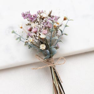 Könnte beinhalten: Ein kleiner Strauß aus Trockenblumen mit violettem Lavendel, weißem Schleierkraut und kleinen rosa Blüten. Die Stiele sind mit einer naturfarbenen Kordel zu einer Schleife gebunden, wobei grüne Eukalyptusblätter in das Gesteck eingearbeitet sind.
