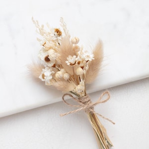 Op de afbeelding: Een klein boeket van gedroogde bloemen in beige en witte tinten, samengebonden met touw. De compositie bevat verschillende grassen en kleine bloemen, wat een natuurlijke, rustieke uitstraling creëert. Ideaal voor bruiloften of huisdecoratie.