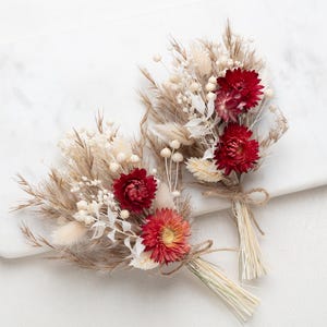 Op de afbeelding: Twee kleine boeketten van gedroogde bloemen, met rode, witte en beige elementen. De boeketten zijn vastgebonden met touw en liggen op een wit marmeren oppervlak. De bloemen omvatten rode dahlia's en diverse gedroogde grassen.