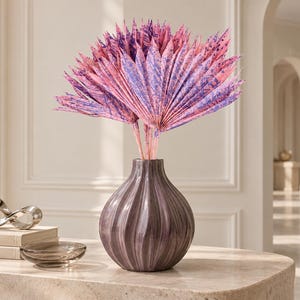 Könnte beinhalten: Ein Strauß aus getrockneten, fächerförmigen Palmblättern in leuchtenden Rosa- und Violetttönen, arrangiert in einer runden, gerippten, dunkelgrauen Keramikvase mit glänzender Oberfläche. Die Palmblätter haben eine markante, gefaltete Struktur.