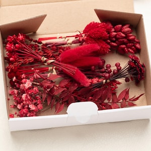 Può includere: Una scatola di cartone piena di vari fiori e fogliame rossi secchi. La composizione include mazzi di piccoli fiori delicati, trame piumate e grappoli simili a bacche. La scatola è aperta, rivelando il contenuto su una superficie bianca.