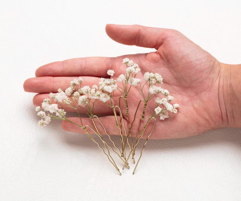 May include: Six gold hair pins with small white dried flowers attached to the ends. The pins are arranged in a hand.