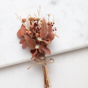 Könnte beinhalten: Ein kleiner Trockenblumenstrauß mit einer Mischung aus Texturen und Farben. Das Arrangement umfasst braune, beige und weiße Elemente, die mit Bindfaden zusammengebunden sind. Ein natürlicher, rustikaler Akzent.
