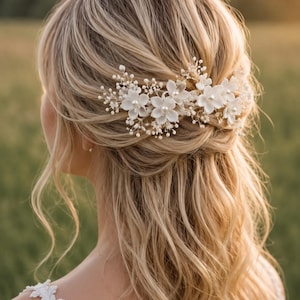 Può includere: Un pettine per capelli con fiori bianchi e perle centrali, intervallato da delicati rametti di piccole perline bianche su filo sottile, fissato in un'acconciatura semiraccolta con capelli biondi ondulati e una spallina in pizzo bianco.