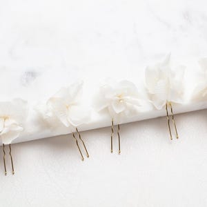 Pode incluir: Cinco grampos de cabelo com flores brancas e hastes de arame dourado. As flores são feitas de tecido e têm uma aparência delicada e com babados.