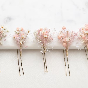 Può includere: Cinque forcine per capelli con piccoli bouquet di fiori secchi nelle tonalità del rosa tenue, crema e bianco. Ogni composizione floreale è fissata con un sottile filo dorato su una forcina a U in metallo color oro.