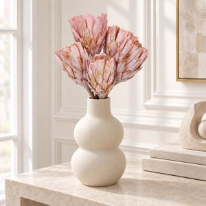Peut inclure: Un bouquet de quatre fleurs de protéa séchées rose clair aux pétales texturés et superposés, disposé dans un vase en céramique blanc cassé, mat et lisse. Le vase présente une silhouette en sablier à double bulbe avec un col étroit.