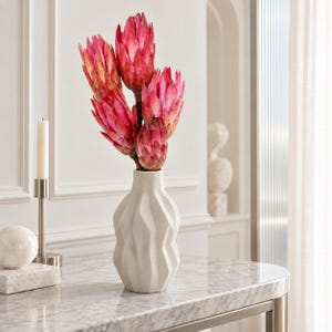 Peut inclure: Un vase en céramique blanche à la surface géométrique et facettée contient un bouquet de cinq fleurs de protéa rose vif. Le vase présente un col étroit et un corps angulaire plus large, révélant une forme sculpturale aux arêtes verticales.