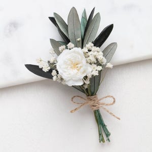 Op de afbeelding: Een kleine, handgemaakte corsage met een witte roos, delicate witte bloemen en olijfbladeren. De bloemstuk is gebonden met touw, wat een rustieke en elegante uitstraling creëert. Perfect voor bruiloften of speciale evenementen.