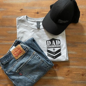 May include: A gray t-shirt with the text "DAD DEDICATED AND DEVOTED" and a black cross and chevron design. A pair of blue jeans and a black baseball cap with the word "Milwaukee" in white text.