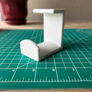 May include: White plastic 3D printed hook with a rounded top. The hook is sitting on a green cutting mat.