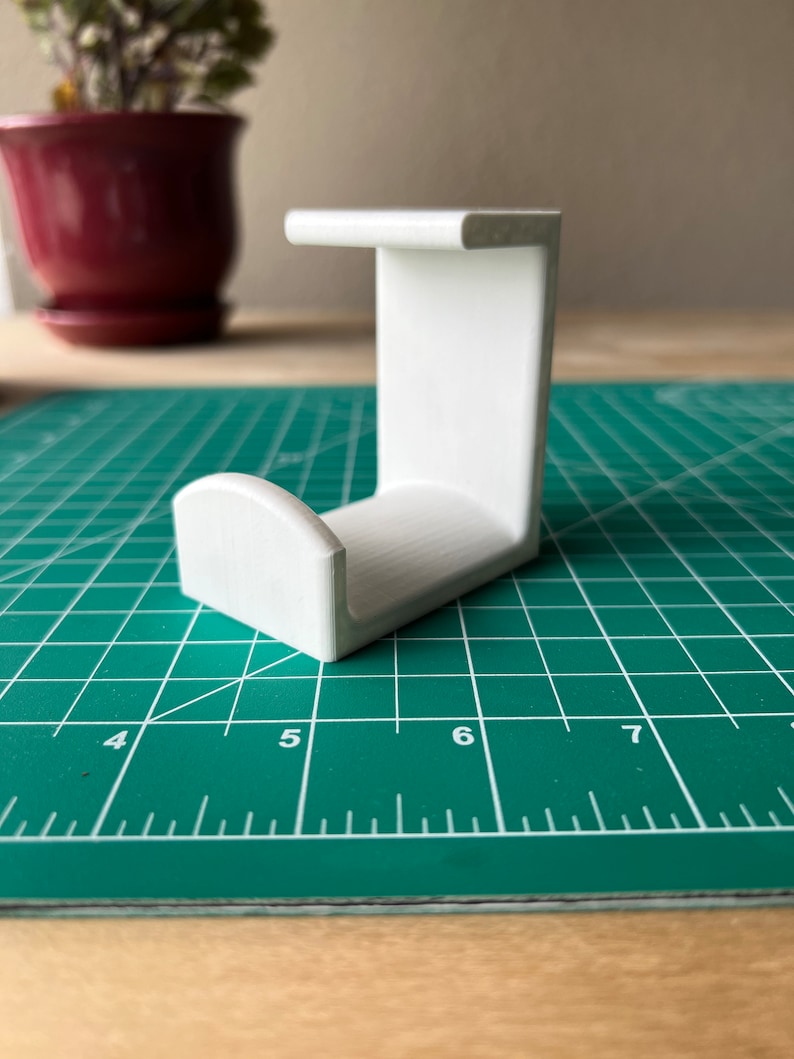 May include: White plastic 3D printed hook with a rounded top. The hook is sitting on a green cutting mat.