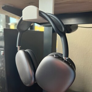 May include: A white plastic headphone hook mounted under a desk, with a pair of black over-ear headphones hanging from it.
