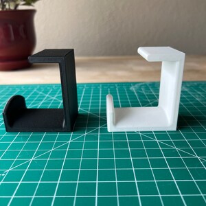 May include: Two 3D printed J-shaped hooks, one black and one white, are displayed on a green cutting mat.