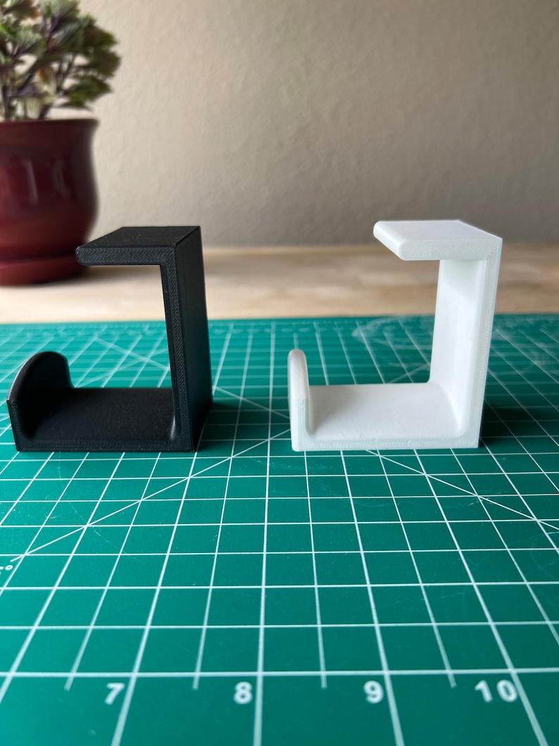 May include: Two 3D printed J-shaped hooks, one black and one white, are displayed on a green cutting mat.
