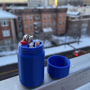 May include: A blue plastic container with a lid, designed to hold a lighter and cigarettes. The container is sitting on a railing with a city skyline in the background.