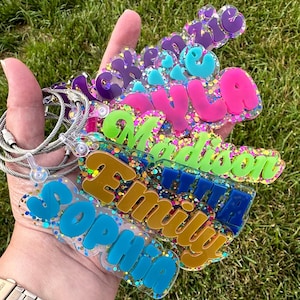 May include: A stack of personalized acrylic keychains featuring names including Mackenzie, Lyla, Madison, Emily, and Sophia. Each keychain has a clear base filled with colorful glitter and a solid-colored raised name in purple, pink, green, gold, or blue.
