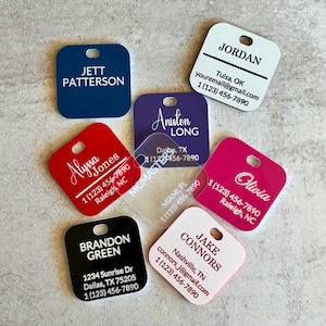 May include: A collection of personalized square tags in various colors, including blue, red, pink, and black. Each tag features a name, contact information, and a small hole for attachment. The tags are designed for identification purposes.