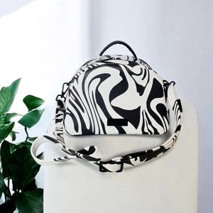 May include: A white and black zebra print handbag with a top handle and a detachable shoulder strap. The bag has a rounded shape and a black buckle. The strap and handle are black.