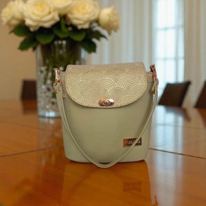 Peut inclure: Un sac à main vert sauge avec un rabat à motifs dorés. Le sac a une bandoulière assortie et des ferrures dorées. Une petite étiquette rectangulaire dorée est sur le devant. Un vase de roses blanches est en arrière-plan.
