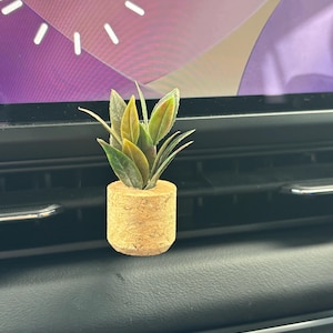 May include: A small artificial plant with green and yellow-tipped leaves housed in a cylindrical, light brown cork-textured base. The plant is positioned against a dark horizontal vent, with a digital clock display visible in the background.