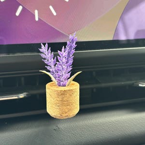 May include: A small car air freshener featuring a cylindrical cork base holding a sprig of artificial purple lavender flowers. The item is designed to be attached to a vehicle's air vent, providing a decorative element for the interior dashboard.