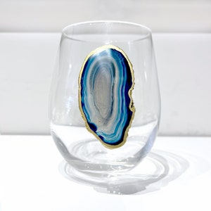 May include: A clear stemless wine glass with a blue, white, and gold agate design. The agate design is oval-shaped and has a gold-colored edge. The glass is sitting on a white surface.