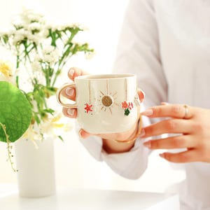May include: Cream-colored ceramic mug with a gold handle and rim. The mug is decorated with a sun and star design, with red and green floral accents. The mug is held in a person's hands.