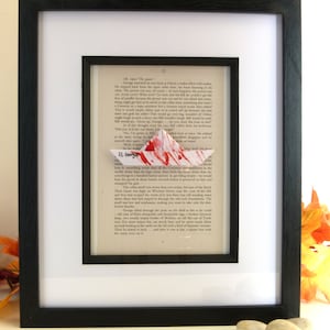 Puede incluir: Obra de arte enmarcada con un diseño de barco de papel con salpicaduras de pintura roja, sobre un fondo de páginas de libros antiguos. El marco negro y el paspartú blanco realzan la obra. El texto "SS. George" es visible.