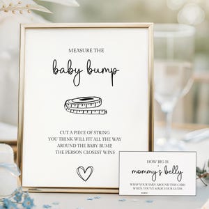 May include: A framed sign with the text "Measure the baby bump" and instructions for a baby shower game. A champagne flute, baby booties, and a small card that says "How big is mommy's belly" are also visible.