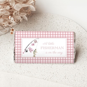 May include: A rectangular candy bar wrapper featuring a pink and white gingham pattern. The center label displays a fishing rod with pink baby clothing hanging from the line, a fish, and the text A little FISHERMAN is on the way.