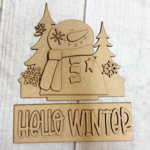 May include: A wooden cutout featuring a snowman with a scarf, flanked by two pine trees and snowflakes. Below, the words "HELLO WINTER" are cut out. The piece is made of unfinished wood.