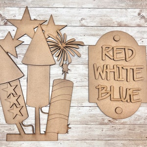 May include: Wooden craft cutouts for a Fourth of July celebration. The set includes three fireworks, two stars, a firework burst, and a sign that reads "RED WHITE BLUE".