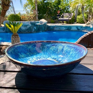 May include: A large, round ceramic bowl with a vibrant blue glaze. The bowl has a dark brown rim and a textured interior, reflecting light. The bowl is sitting on a dark wooden surface, with a pool and greenery in the background.