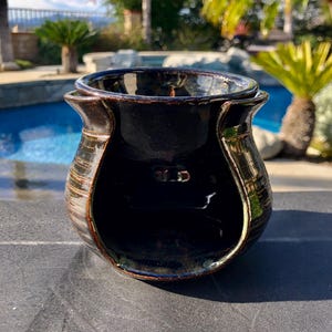 May include: A dark brown and black glazed ceramic wax warmer with a glossy finish. The vessel features a wide, rounded body with a large arched opening at the front and a separate, removable dish resting on the top rim for holding wax melts.
