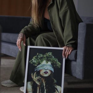 Puede incluir: Una impresión artística enmarcada que presenta a una mujer con una venda en los ojos y una corona floral. La obra de arte tiene un fondo oscuro y los labios de la mujer son de un rojo intenso. El marco es blanco.