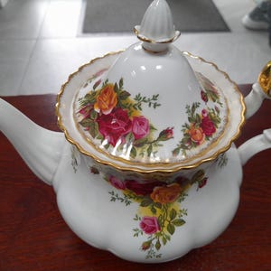 Peut inclure: Théière en céramique blanche avec couvercle, ornée de roses rouges, oranges et roses. La théière a une bordure dorée sur le couvercle et la poignée. Le bec et la poignée sont blancs. Un article de service à thé classique.