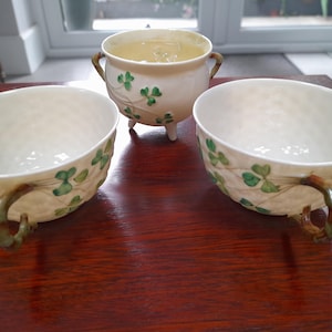 Puede incluir: Juego de tres tazas de té de cerámica color crema y un cuenco pequeño en forma de caldero. Cada pieza está decorada con diseños de tréboles verdes y asas marrones. El caldero tiene tres patas pequeñas y dos asas.